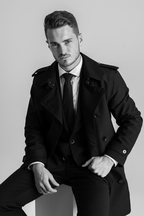 Andreas Schmid, black and white photo, seated in a trench coat and tie
